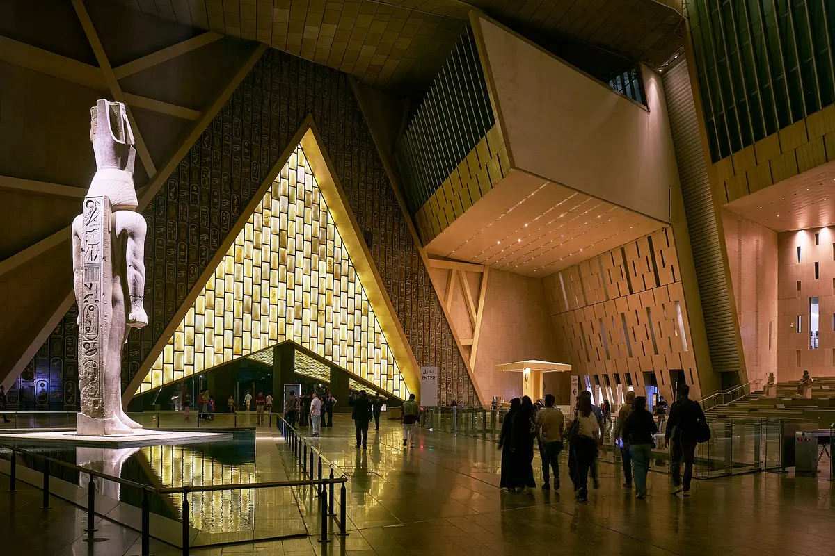 The standing statue of Ramesses II beside a reflecting pool in the Grand Hall, with the lit triangular entrance glass behind it