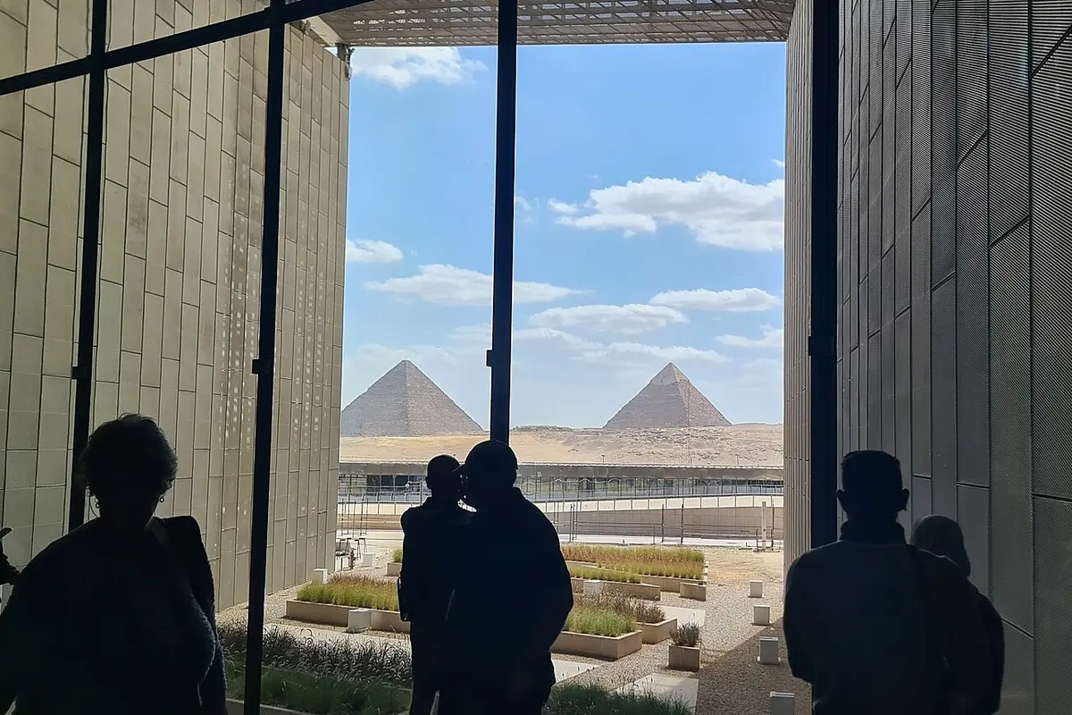Two of the Giza pyramids seen across the museum's garden, framed by an opening in its walls, with visitors in silhouette in the foreground
